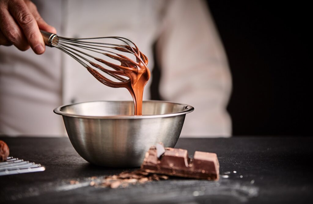 Szef kuchni ubija roztopioną czekoladę w misce ze stali nierdzewnej za pomocą drucianej trzepaczki z bliska na dłoni