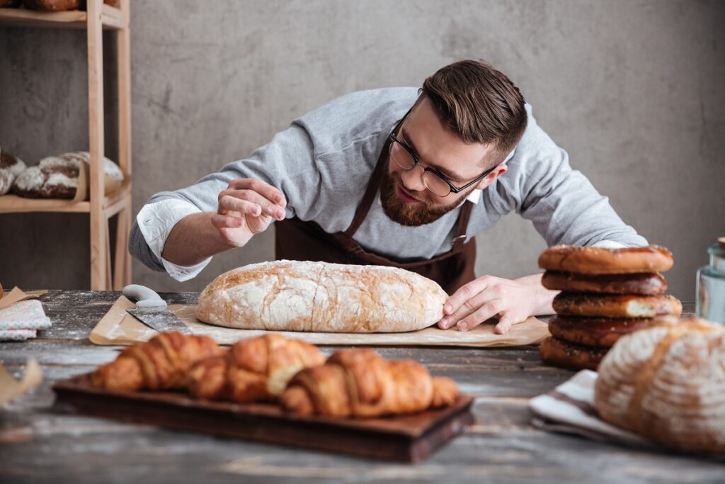 Piekarz pochyla się nad bochenkiem chleba i posypuje go mąką