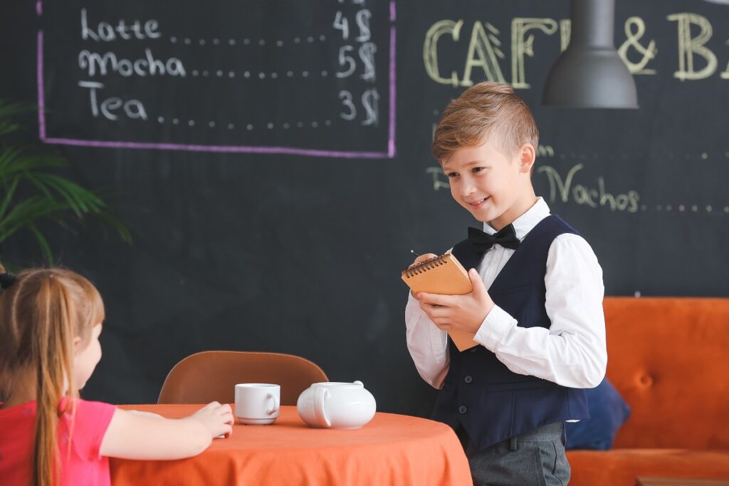 Chłopiec jako kelner zapisuje zamówienie od dziewczynki, która siedzi przy stoliku