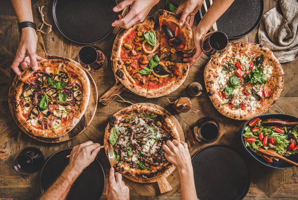 Różnego rodzaju pizze na stole i ręce, które po nie sięgają. 